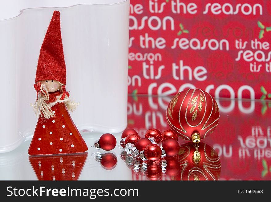 Red christmas balls on mirror red background reading tis the season and red christmas fairy standing on front of white backdrop. Red christmas balls on mirror red background reading tis the season and red christmas fairy standing on front of white backdrop