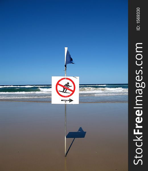 A no surfing sign is displayed at the beach. The sky is clear blue. Note : The sign is naturally weathered.