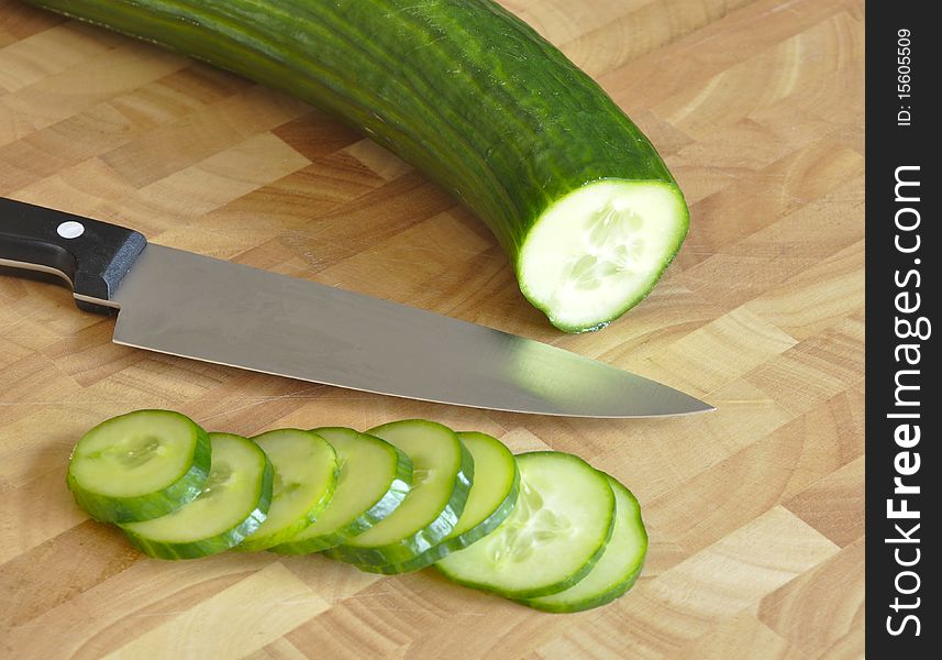Kitchen knife rests on wooden chopping board with sliced cucumber.