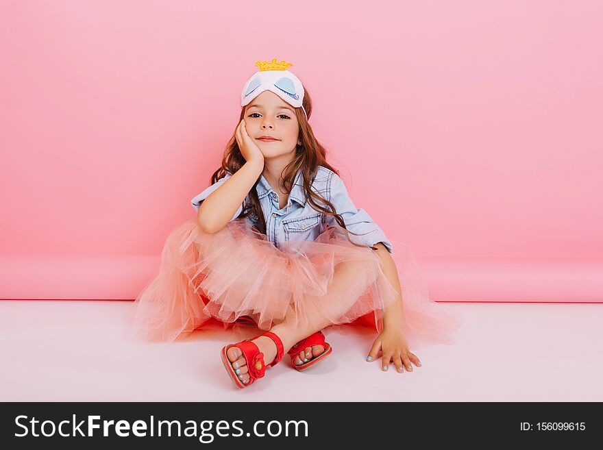 Joyful Little Girl In Tulle Skirt Sitting On White Floor Isolated On Pink Background. Pretty Princess Child With Mask On