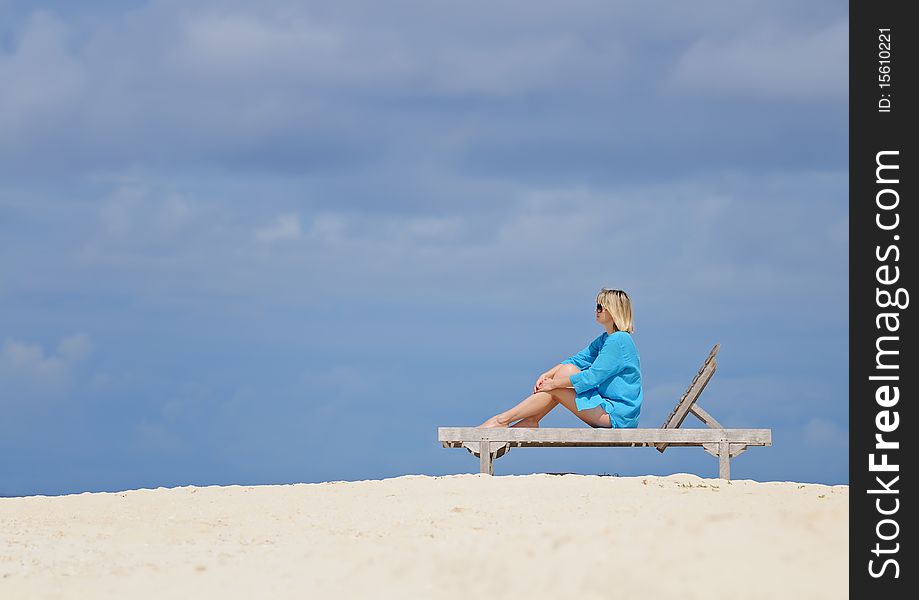 Woman in blue tunic sitting on wooden deck-chair on sandy beach against cloudy sky. Woman in blue tunic sitting on wooden deck-chair on sandy beach against cloudy sky.