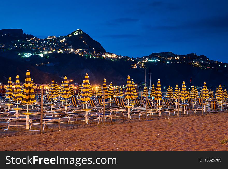 Beach at night with an illuminated village in the mountains and blue sky. The name of the village is Taormina, which is situated on Sicily, Italy.