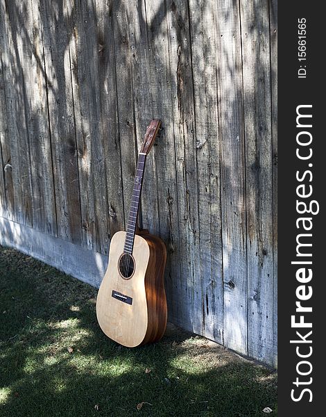 Its an shaded, custom, acoustic guitar viewed from an angle, placed against a faded fence resting on a bed of green grass. Its an shaded, custom, acoustic guitar viewed from an angle, placed against a faded fence resting on a bed of green grass.