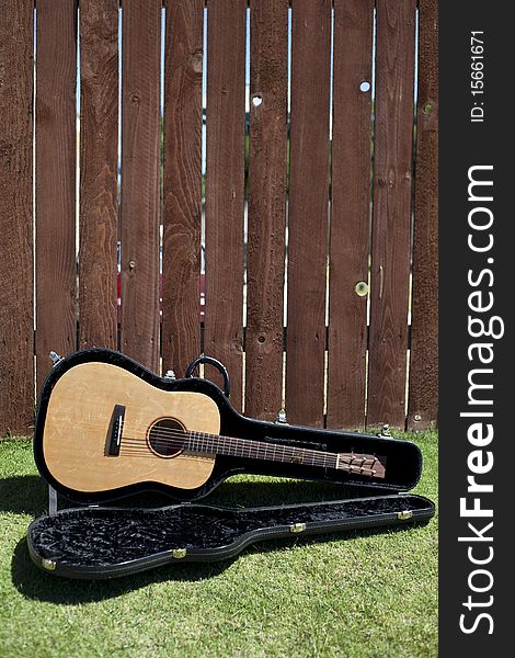 This is a picture of an acoustic guitar in a case against a redwood fence with a long view of the fence. This is a picture of an acoustic guitar in a case against a redwood fence with a long view of the fence..