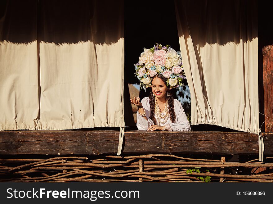 Brunette girl in a white ukrainian authentic national costume and a wreath of flowers is is looking out of the window.