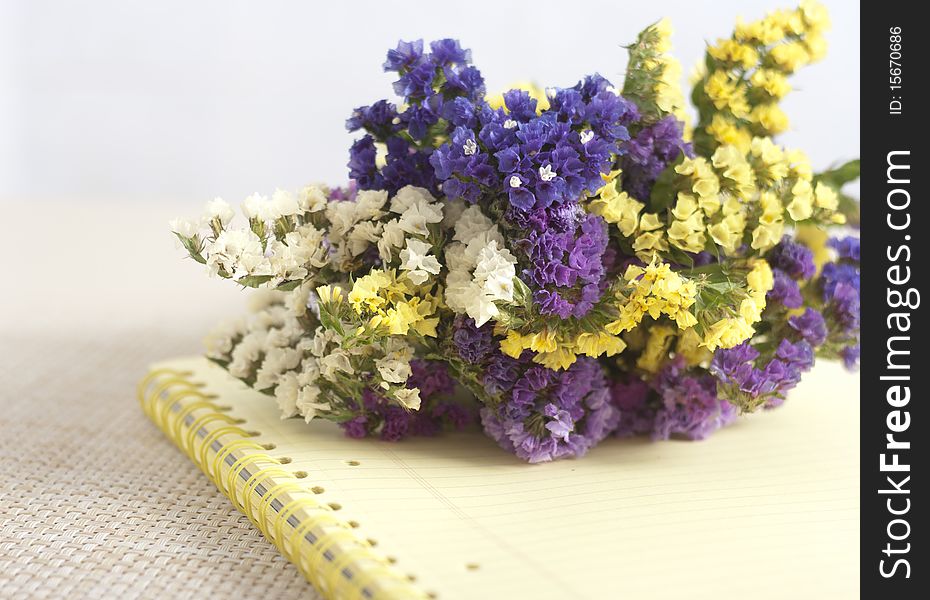 A bunch of violet,blue, yellow and white immortelles on a yellow copybook lying on a straw tabletop. A bunch of violet,blue, yellow and white immortelles on a yellow copybook lying on a straw tabletop