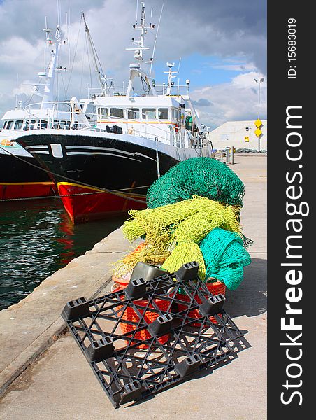 Green and blue-green fishing nets stacked along with with a crate and buckets on a quayside in Ireland. The nets are pictured in front of a trawler with a black and red hull. Green and blue-green fishing nets stacked along with with a crate and buckets on a quayside in Ireland. The nets are pictured in front of a trawler with a black and red hull.