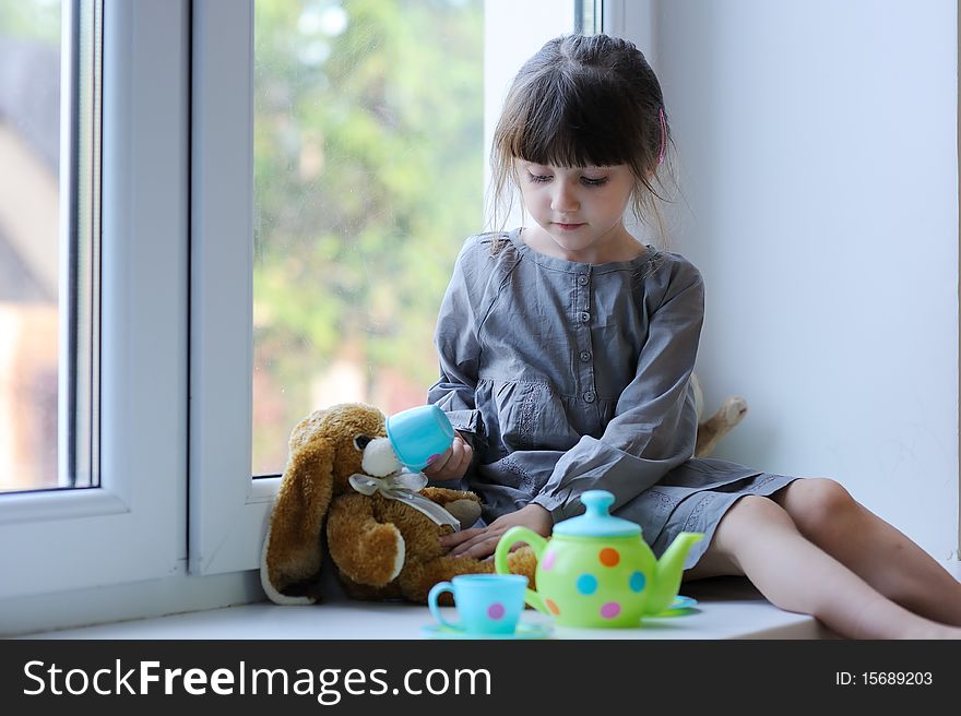 Nice toddler girl with toy tea set and bunny sitting near the winow in cute gray dress