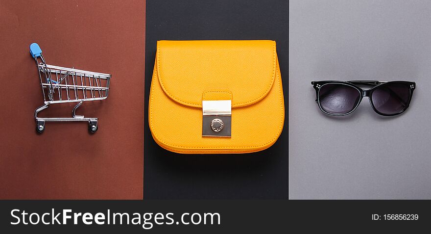 Minimalistic Shopaholic Concept. Yellow leather bag, mini shopping trolley sunglasses on paper background. Women&#x27;s fashion accessories. Top view