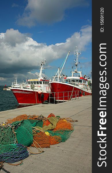 Green and blue and orange fishing nets stacked on a quayside in Ireland. The nets are pictured in front red and white trawlers. The sky is a rich blue with cumulus clouds. Green and blue and orange fishing nets stacked on a quayside in Ireland. The nets are pictured in front red and white trawlers. The sky is a rich blue with cumulus clouds.