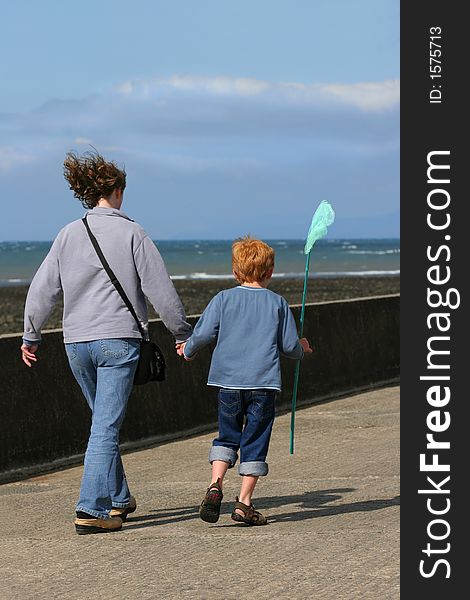 Woman holding the hand of a young boy holding a green fishing net in his hand and walking along a beach promenade. Sea out of focus to the rear. Woman holding the hand of a young boy holding a green fishing net in his hand and walking along a beach promenade. Sea out of focus to the rear.