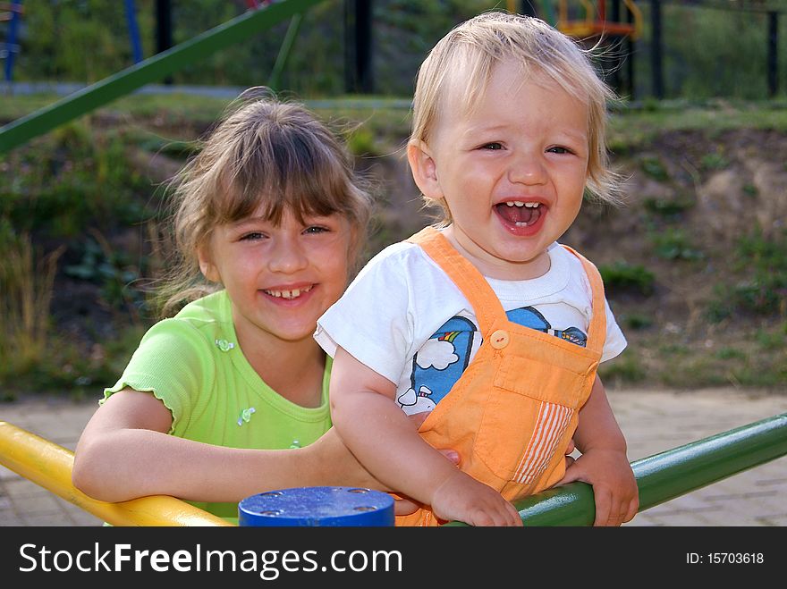 In the summer on a children's playground the girl smiles and holds the younger brother who shouts. In the summer on a children's playground the girl smiles and holds the younger brother who shouts