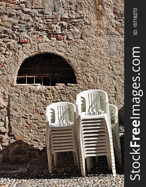 Some stacks of modern white plastic chairs with an old wall like background in a street of Garessio, in Piedmont, Italy.