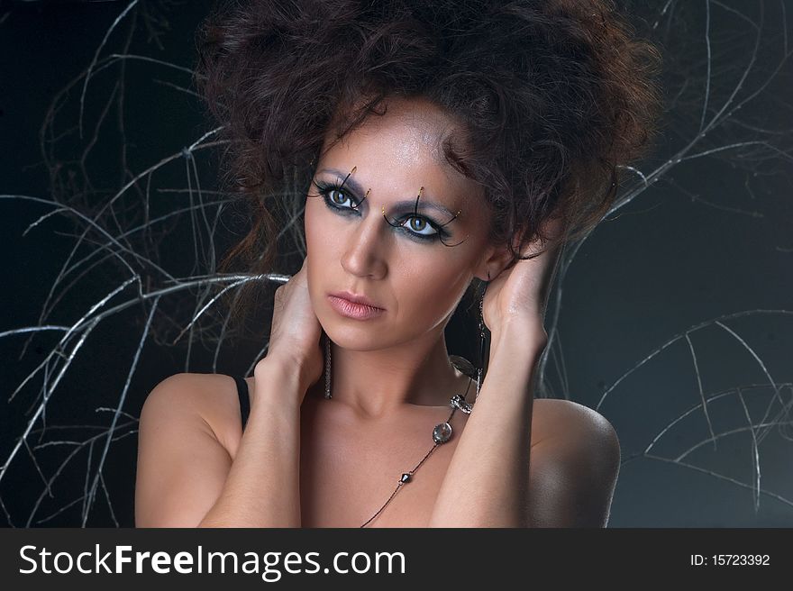 Portrait of a lovely woman in a bizarre makeup. The image is taken on a dark and mystique background with branches.