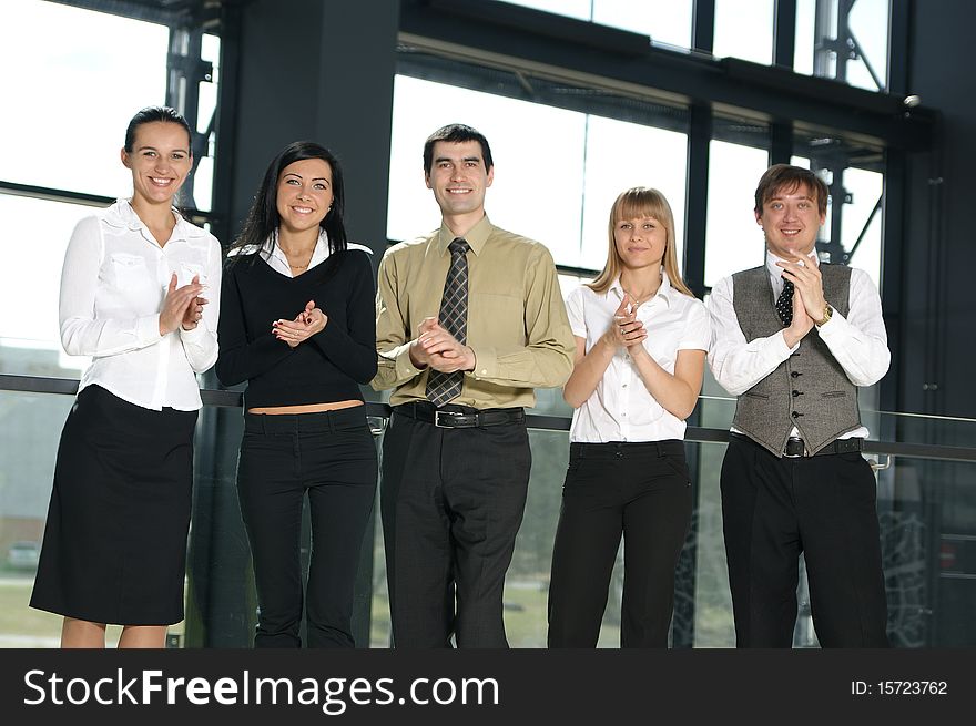 Five business persons are clapping their hands together. The image is taken in a modern office.