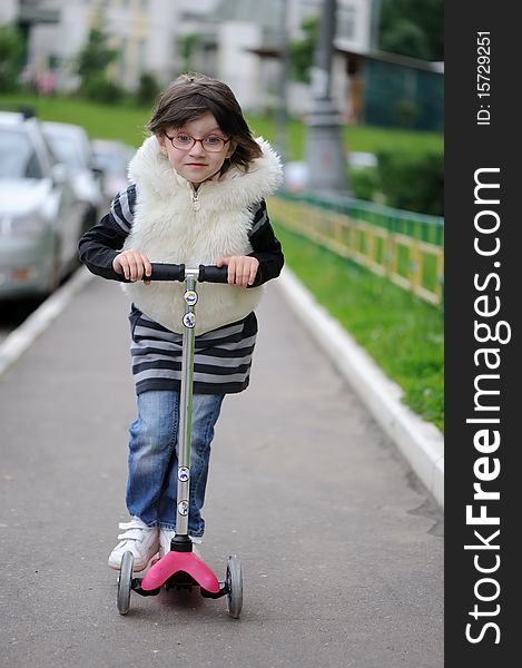 Nice little toddler girl in stripe dress, jeans and white fur jacket riding on her pink scooter. Nice little toddler girl in stripe dress, jeans and white fur jacket riding on her pink scooter