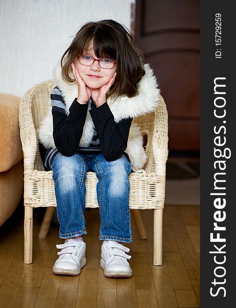 Nice toddler girl in glasses and white fur jacket with dark hair sitting in chair and looking to the camera. Nice toddler girl in glasses and white fur jacket with dark hair sitting in chair and looking to the camera