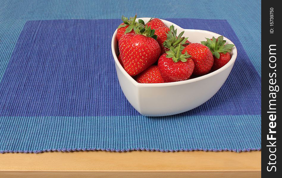 Strawberries in a heart shaped bowl on a blue place mat on a breakfast table