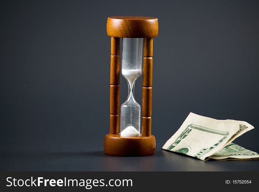 Folded twenty dollar bill is seen next to a wooden hourglass against black background. This image is  a metaphor for the saying  Time is Money