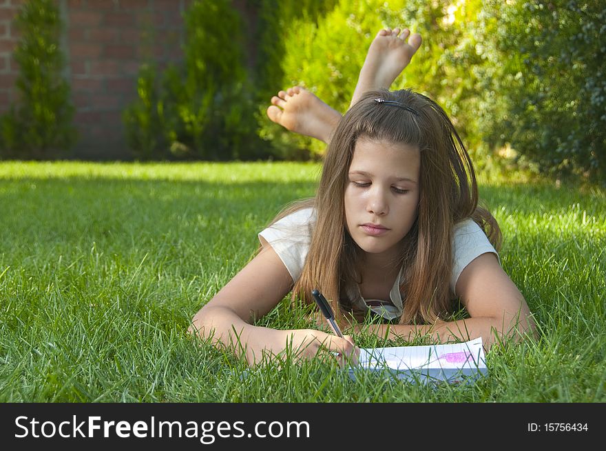 Pretty young teenage girl drawing on the paper while lying on the bed of grass.