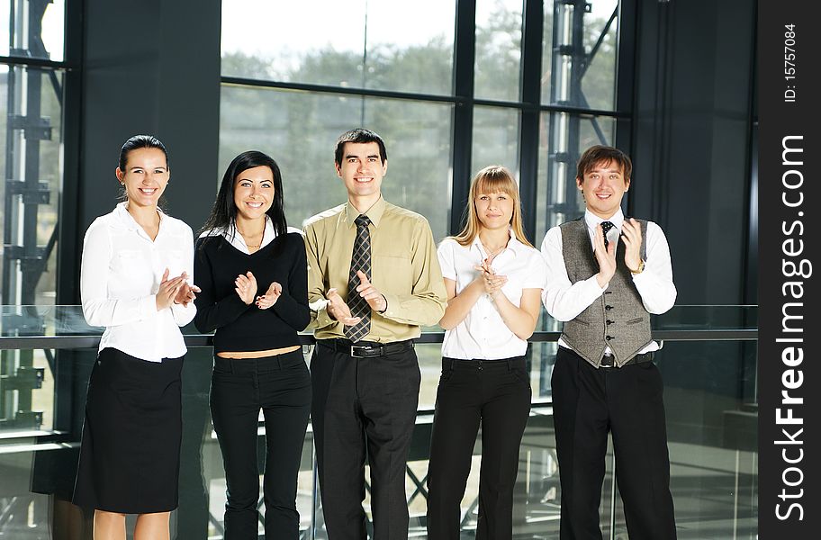 A group of five young business persons is clapping their hands. The image is taken on a modern office background. A group of five young business persons is clapping their hands. The image is taken on a modern office background.