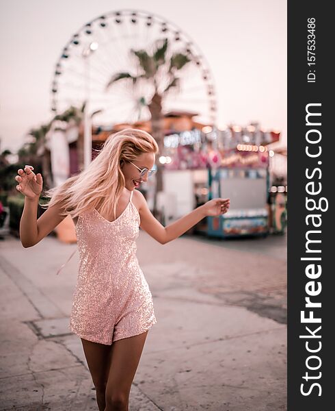 Beautiful hipster girl in stylish fashion dress and jacket in glamour blue sunglasses happy posing at the Ferris wheel near Agia Napa amusement park carousel. Beautiful hipster girl in stylish fashion dress and jacket in glamour blue sunglasses happy posing at the Ferris wheel near Agia Napa amusement park carousel