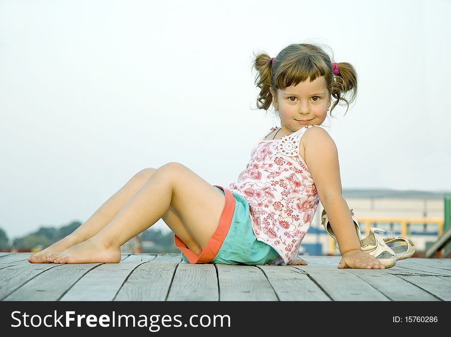 The child sits in the summer in a dress on the bridge sideways and looks aside. The child sits in the summer in a dress on the bridge sideways and looks aside