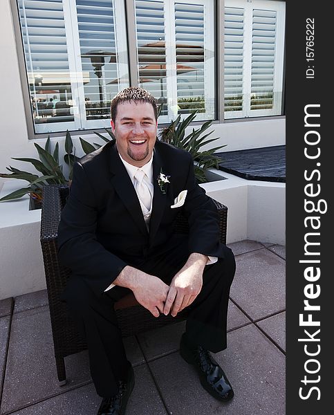 A handsome groom wearing black tuxedo laughing sitting on patio.