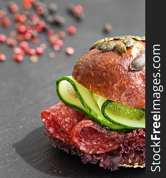 Burger with salami, lettuce and cucumber. Bun with sesame seeds and pumpkin seeds. Quick breakfast. Black stone background. Close-up