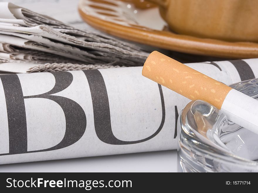 A filtered cigarette resting on a glass ashtray next to a brown cup and plate and a newspaper.