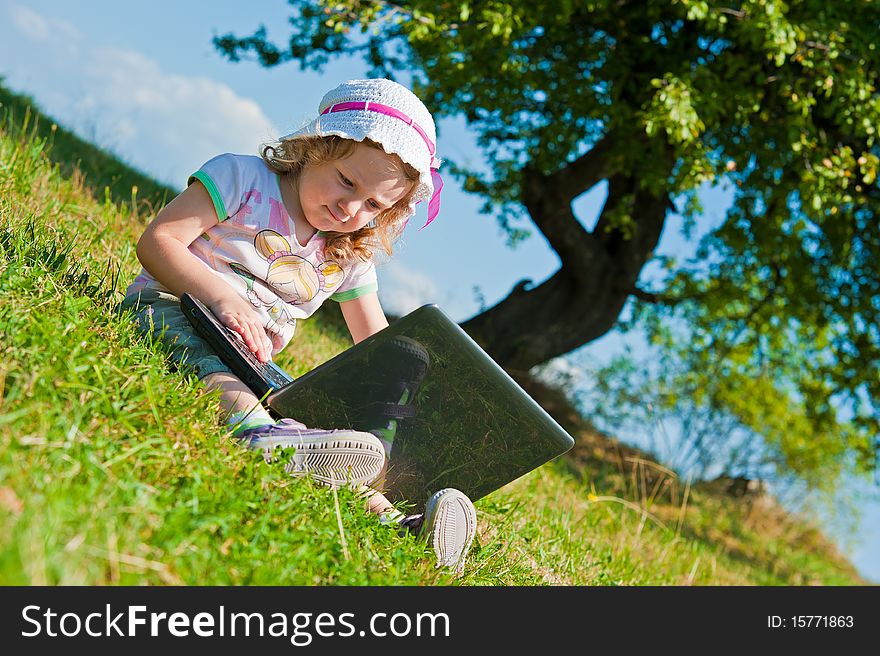 Portrait of a little girl, using notebook computer in the middle of the field, sitting on the grass, making her first steps in technology. Portrait of a little girl, using notebook computer in the middle of the field, sitting on the grass, making her first steps in technology