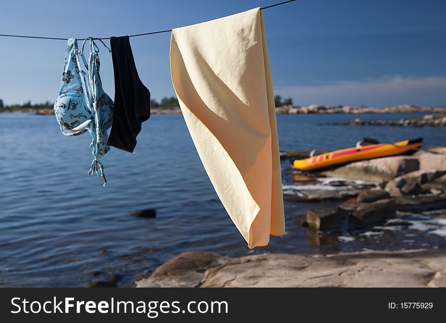 Women's and men's swimsuits and a towel drying on a laundry line at a lake with a kayak on a rocky beach. Women's and men's swimsuits and a towel drying on a laundry line at a lake with a kayak on a rocky beach.