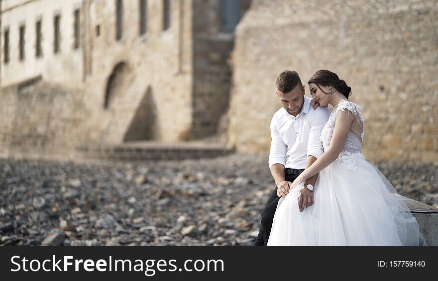 Close-up of beautiful fairytale newlywed couple sitting on the bench and hugging at rocky beach near old medieval castle
