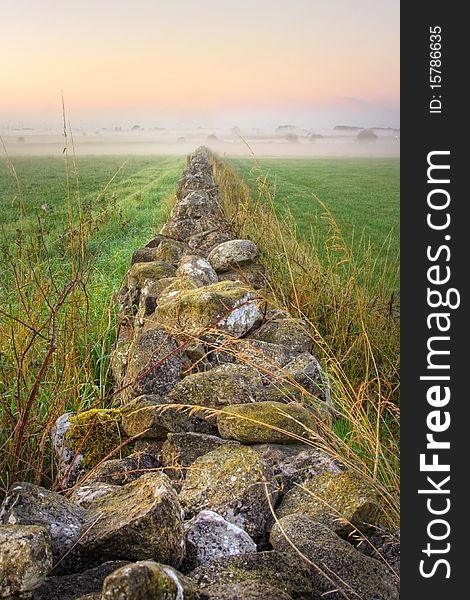 Sunrise at field with stone wall vanishing in the fog and silhouette of trees, HDR image. Sunrise at field with stone wall vanishing in the fog and silhouette of trees, HDR image