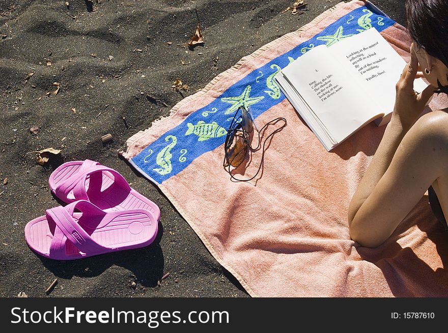 Beach scene with a partial view of a woman lying on an orange beach towel reading a book in the right side and a pair of pink sandals on the left; horizontal view