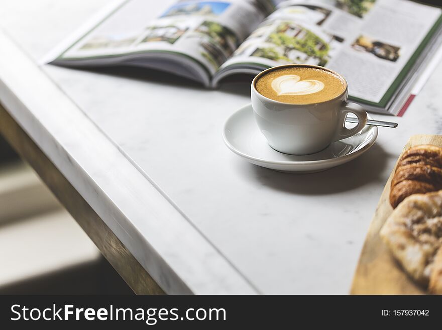 A cup of cappuccino with a cute saucer heart, a croissant, and a magazine on the white table