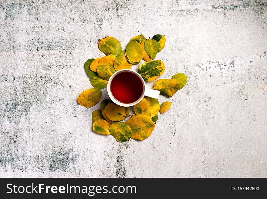 Cup of hot tea decorated by yellow autumn leaves over grey texture background