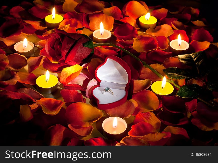 The professions of love represented by the rose petals with ring in box and red rose lit up by candles on the table.