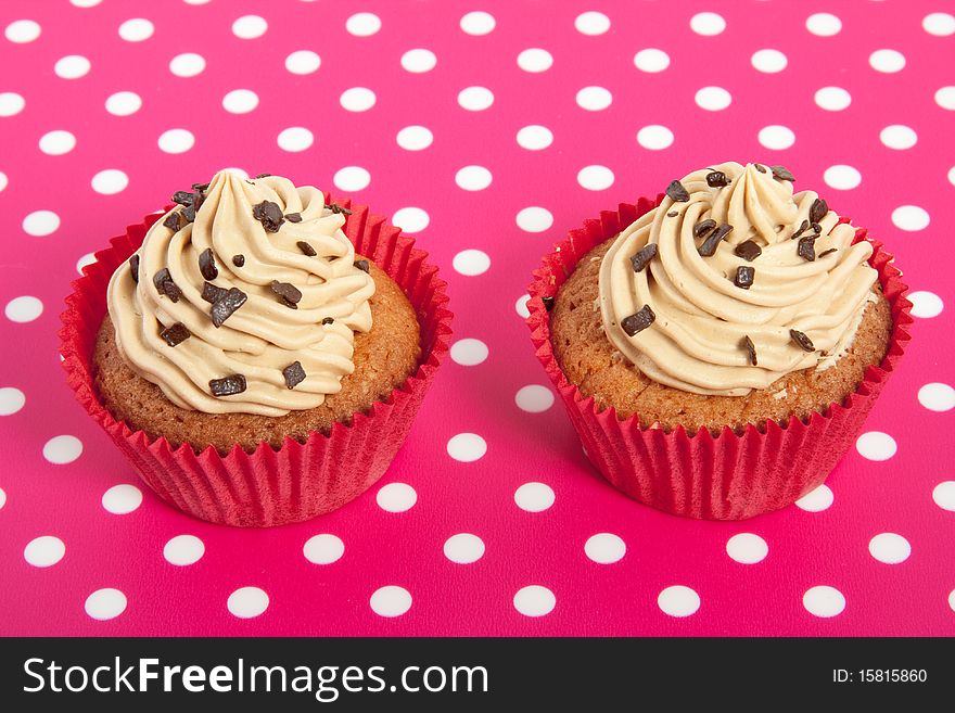 Two red cupakes with cream and chocolate on a pink white dotted background. Two red cupakes with cream and chocolate on a pink white dotted background