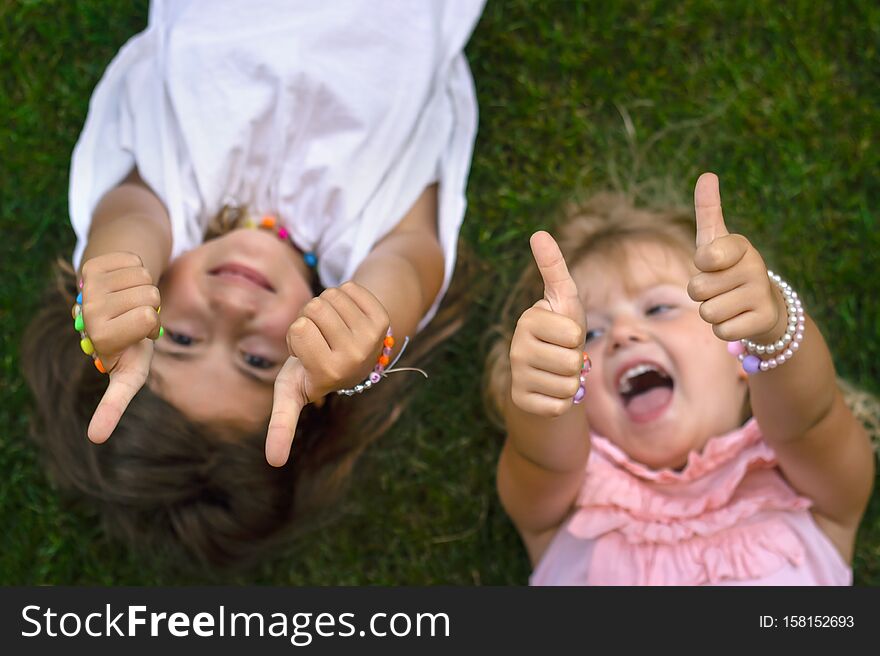 Two Little Girls Laying On The Grass, Laughing And Showing Thumbs Up