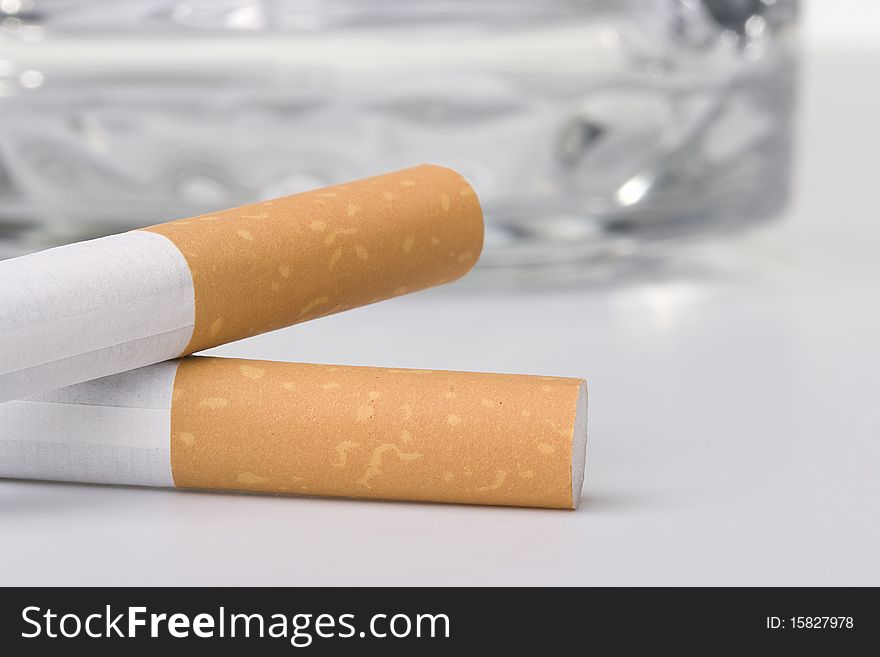Two filtered cigarettes laying in front of a glass ashtray.