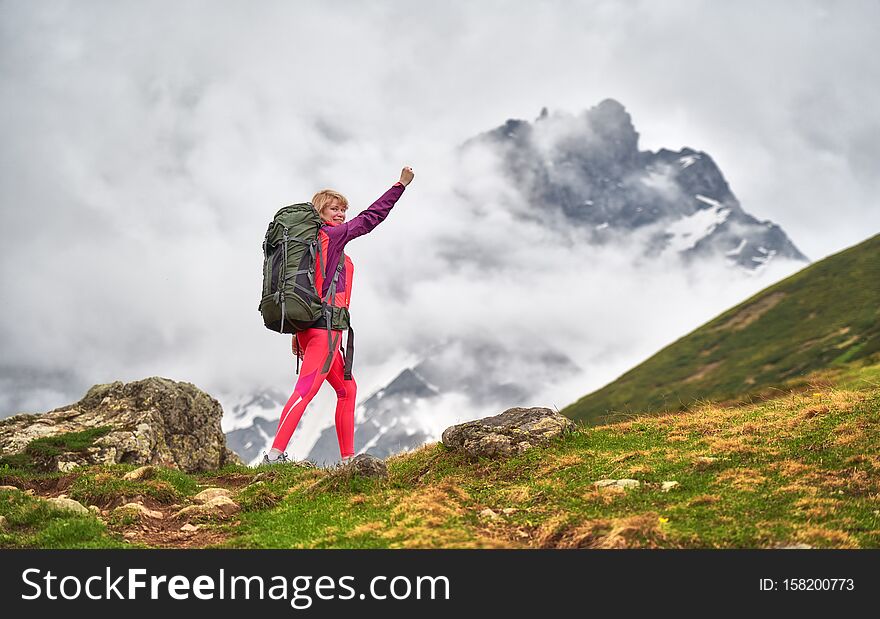Portrait of blonde hiking woman in red clothes and sunglasses holding her arm up at the beautiful mountains background. cloudy weather. Portrait of blonde hiking woman in red clothes and sunglasses holding her arm up at the beautiful mountains background. cloudy weather