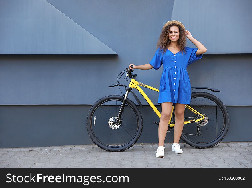 Beautiful young African-American woman with bicycle near grey wall on city street. Space for text