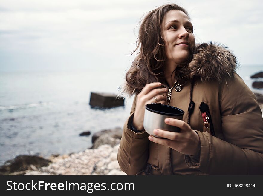 Smiling beautiful young woman standing on sea coast during cloudy weather with traveler cup in hands