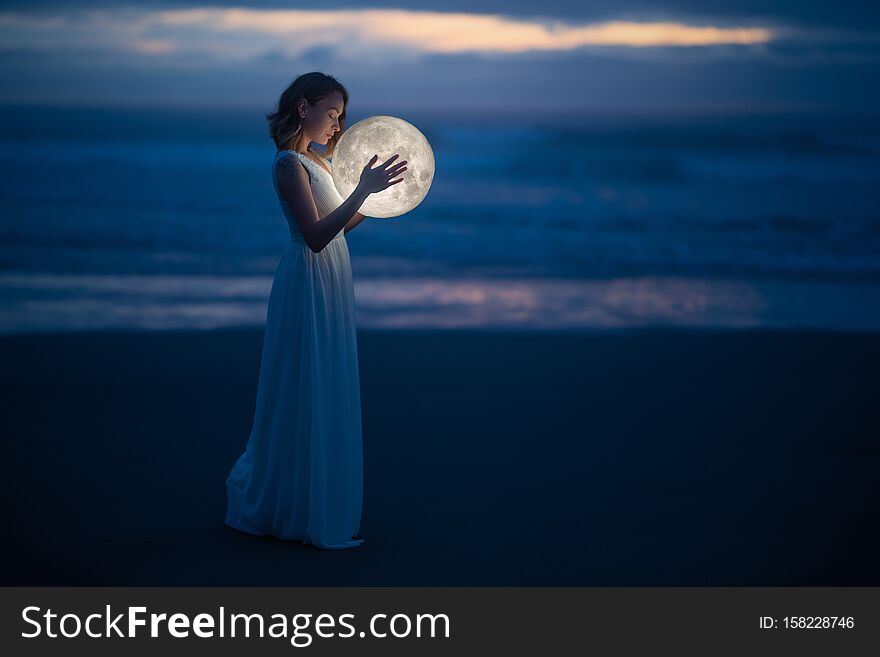 Tender image of a girl. Female magic. Beautiful attractive girl in full growth on a night beach with sand hugs the moon, art photo