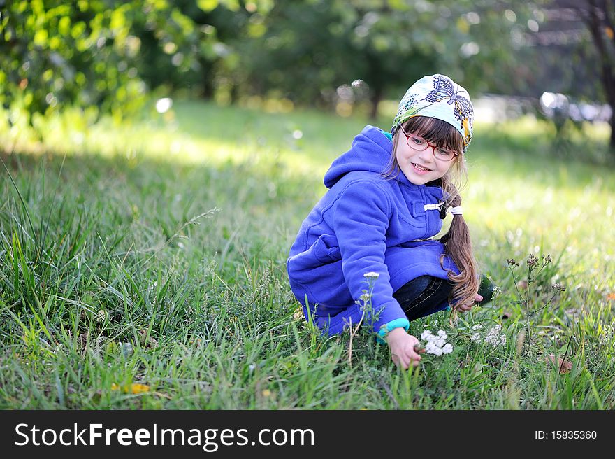 Nice playful little girl in blue coat and glasses picking up flowers on green background. Nice playful little girl in blue coat and glasses picking up flowers on green background