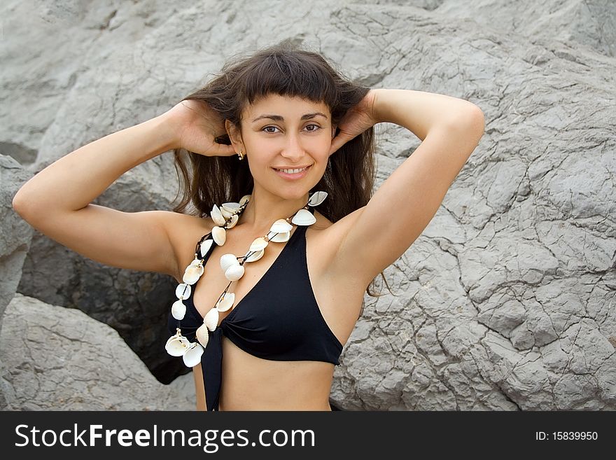 Young pretty girl in the bathing suit with seashell beads near the rocks on the seashore. Young pretty girl in the bathing suit with seashell beads near the rocks on the seashore