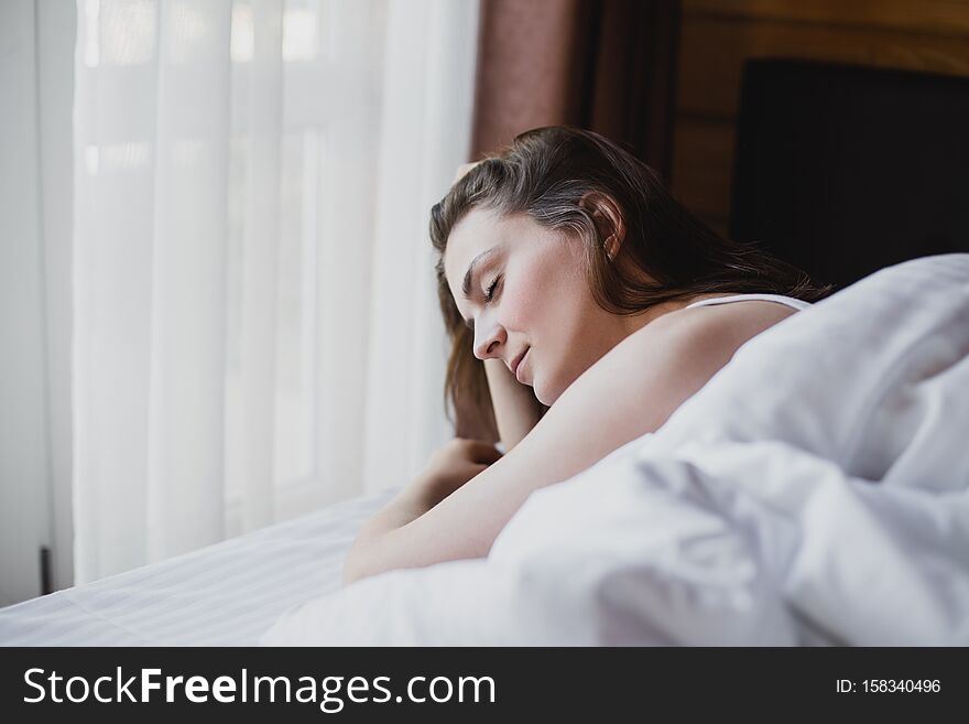 Beautiful young woman having good morning in bed with clean white linens in a cozy wooden house. She is looking at window and having dreamy mood. Beautiful young woman having good morning in bed with clean white linens in a cozy wooden house. She is looking at window and having dreamy mood.