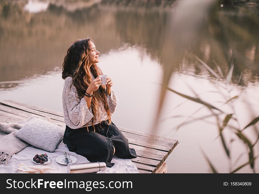 Happy woman in knitted sweater drinking tea near lake.