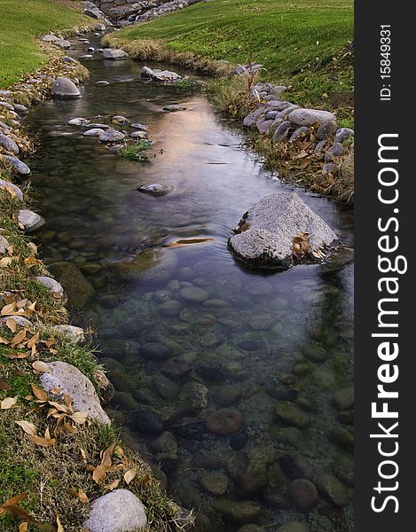 A slow moving creek with green grass and fall leaves glows as the sun rises. A slow moving creek with green grass and fall leaves glows as the sun rises.